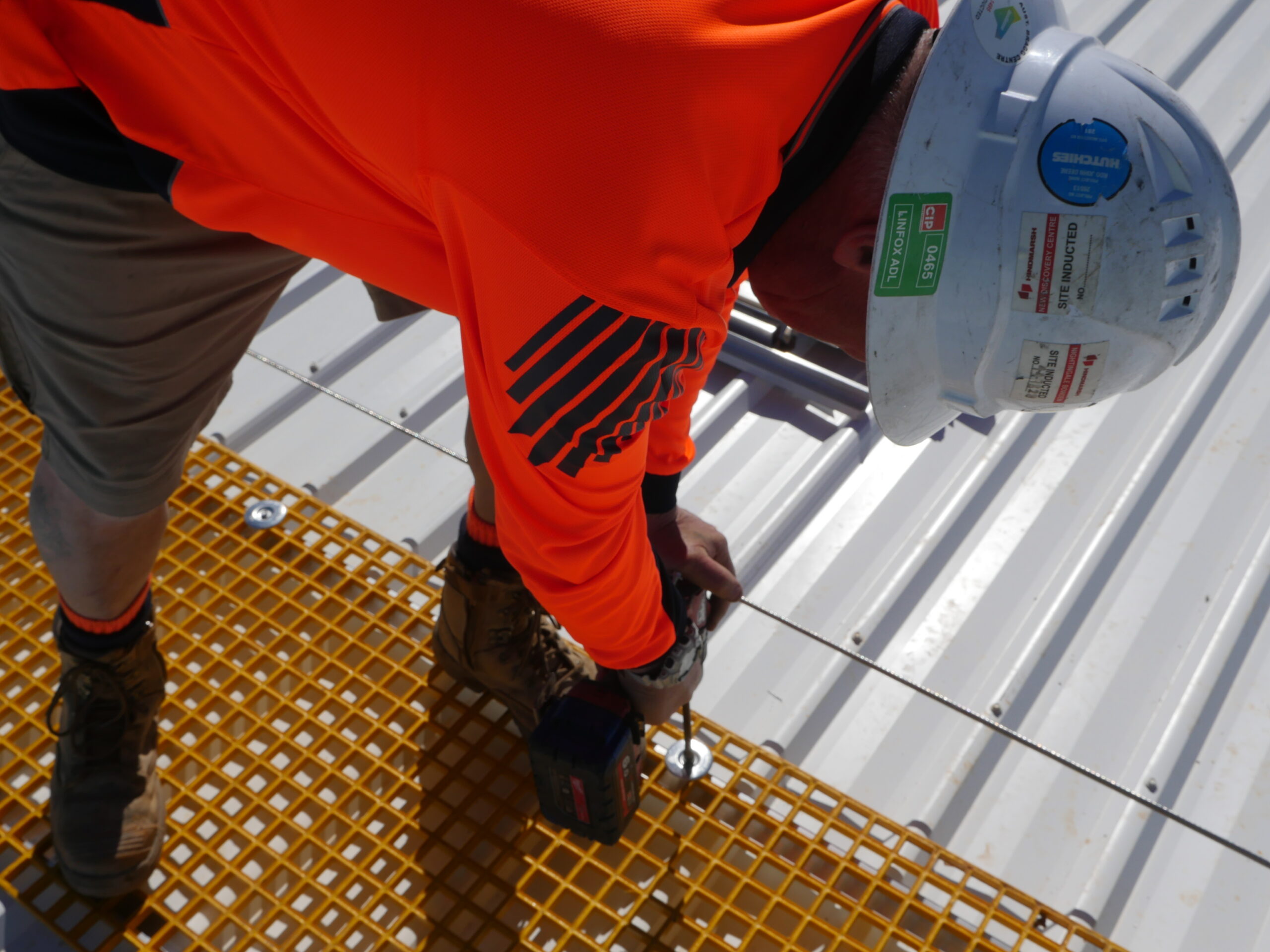 Worker installing safety equipment on a yellow safety grid atop a metal roof, wearing an orange high-visibility shirt and a safety helmet, emphasising RISSafety's commitment to fall prevention systems.