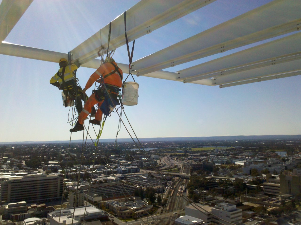 Two workers in safety harnesses performing maintenance on a high-rise building, showcasing RISSafety's fall prevention systems in action, with a panoramic view of the city below.