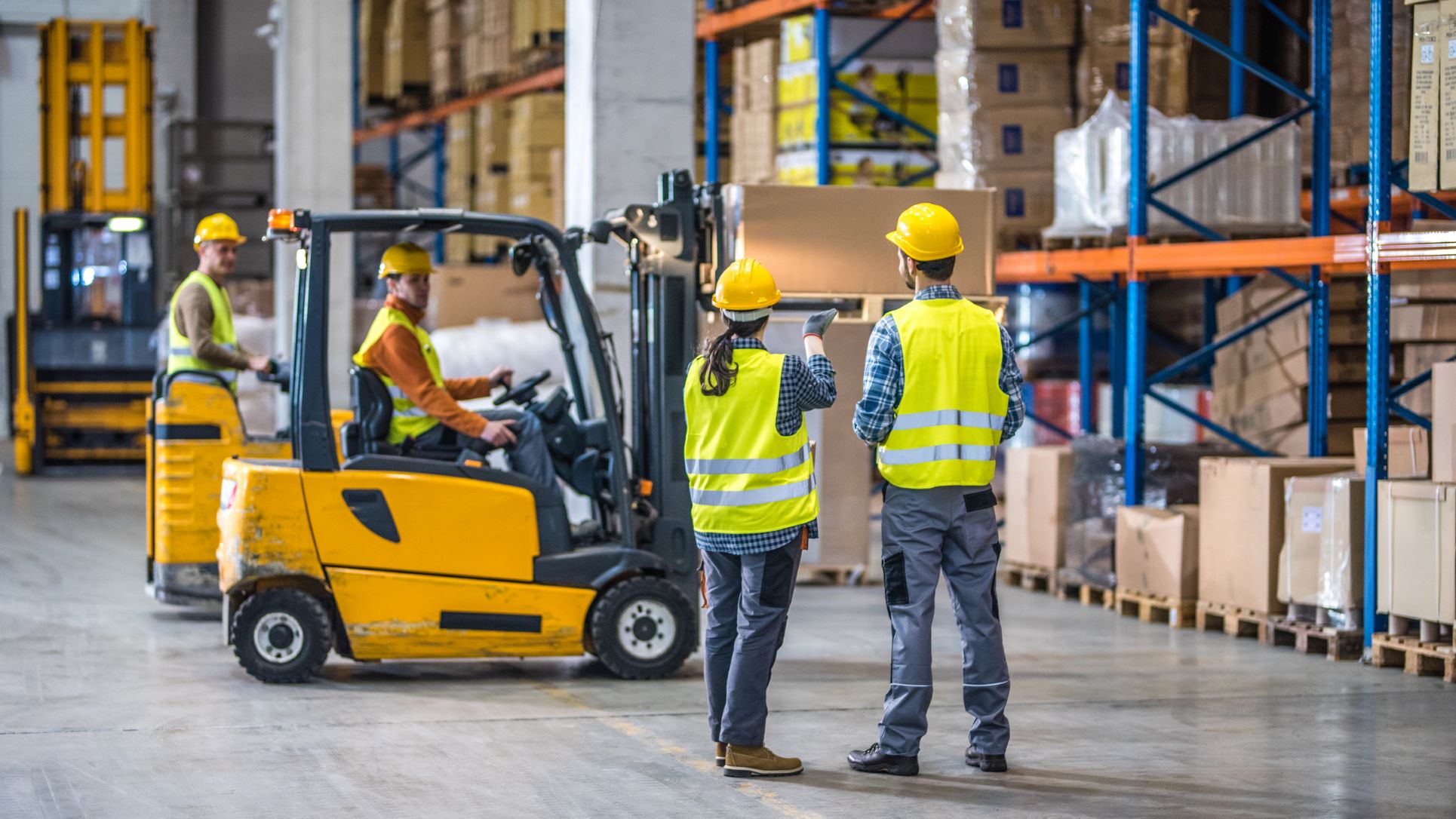 Workers in high-vis vests and hard hats discussing workplace safety in a warehouse environment.