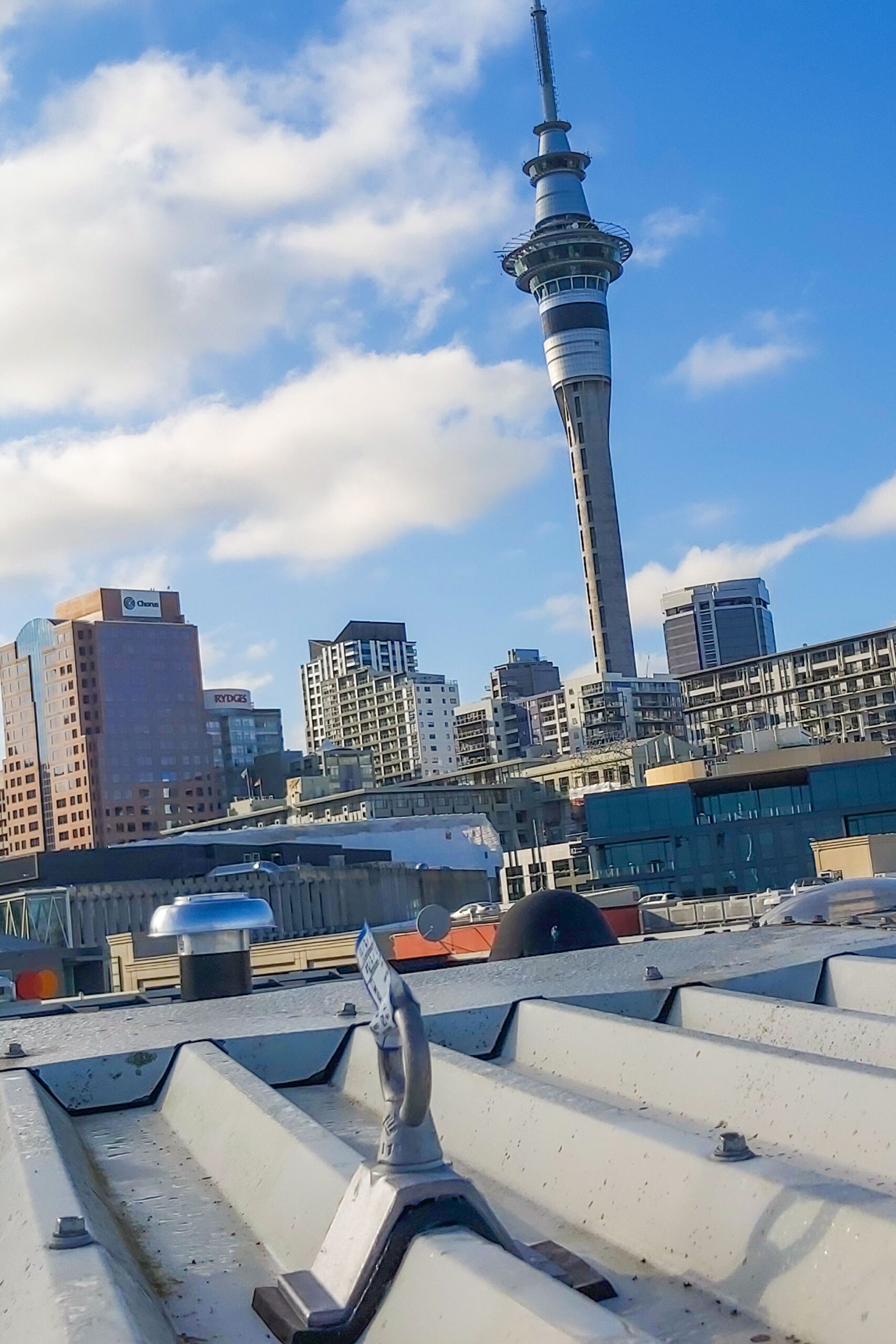 Industrial rope access system anchor point on a rooftop with Auckland skyline and Sky Tower in the background.